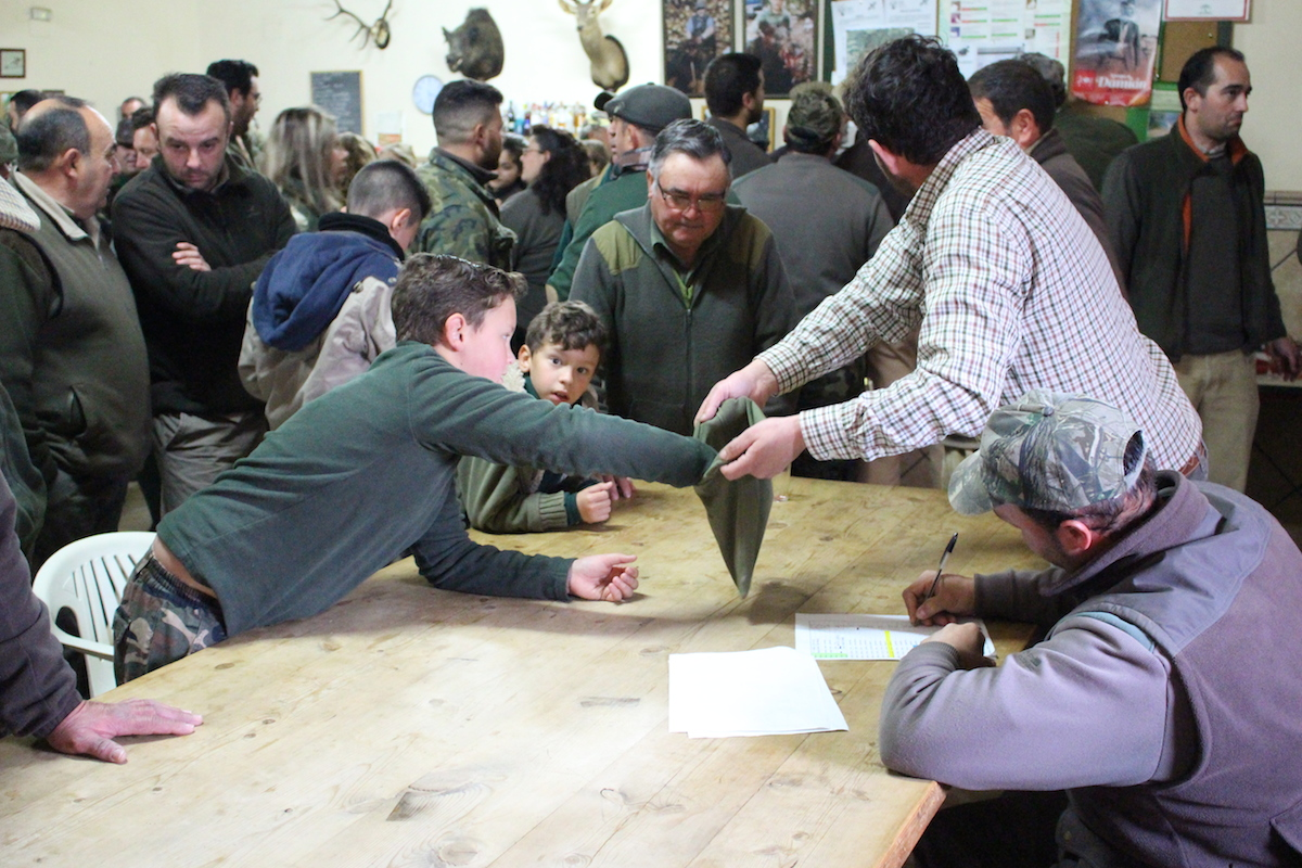 El sorteo (sede de la Sociedad de Cazadores en Paterna del Campo, Huelva)
La monter&iacute;a tiene un ritual con una serie de actos secuenciados, el primero de los cuales es el sorteo. Mediante este acto, los cazadores ser&aacute;n ubicados en los distintos &ldquo;puestos&rdquo; o &ldquo;puertas&rdquo; de las &ldquo;armadas&rdquo; (serie sucesiva de puestos dentro de la &ldquo;mancha&rdquo; o territorio de caza). Ser&aacute; la suerte la que dirimir&aacute; las posiciones en las que, eventualmente, entrar&aacute;n las reses. Despu&eacute;s, ser&aacute; la pericia de cada cazador la que garantizar&aacute; que se consiga o no abatir al animal.
Los sorteos se realizan tanto en el campo, como en las sedes de las sociedades de cazadores. En este caso, los socios han acudido temprano para desayunar (migas, tostadas y caf&eacute;, an&iacute;s), haciendo del momento previo a la salida al campo un espacio ritualizado de encuentro y concentraci&oacute;n de la expectativa de la partida de caza. El sorteo se escenifica desde una mesa alargada desde donde el secretario de la sociedad va llamando a cada socio para que extraiga la papeleta que le ubicar&aacute; en la puerta correspondiente. A continuaci&oacute;n, el presidente dar&aacute; unas indicaciones sobre seguridad, las especies que pueden ser capturadas, si es posible descastar hembras o no, normas de comportamiento del buen cazador o sobre la comida con que se cerrar&aacute; la jornada.
A pesar de lo temprano de todo este proceso, los ni&ntilde;os imitan a sus mayores: se visten de monter&iacute;a, acuden al sorteo y van conociendo los tiempos y ritmos de la actividad. S&oacute;lo algunos de ellos acompa&ntilde;ar&aacute;n, pasados unos a&ntilde;os, a alg&uacute;n familiar o amigo a los puestos desde donde aprender en la pr&aacute;ctica el h&aacute;bito de montear.